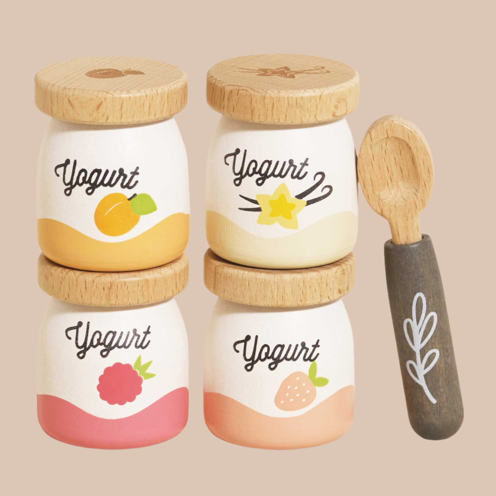 The Yogurt Play Food Pack includes four small jars with wooden lids and fruit icons: peach, star fruit, raspberry, and strawberry. It comes with a wooden spoon featuring a leaf design, ideal for imaginative play on a soft beige background.