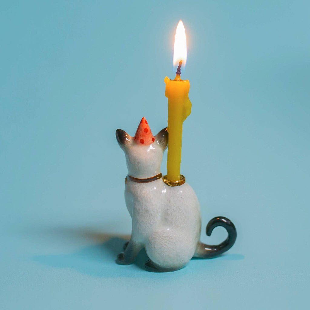 The Siamese Cat Cake Topper, a whimsical ceramic figurine with a red party hat and lit yellow candle, set against a light blue background, adds festive charm to any cat-themed party.