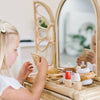 A blond-haired child, absorbed in play at a wicker vanity table, holds an item from a non-toxic painted Wooden Beauty Kit, creating a timeless scene against a softly blurred background.