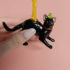 A hand holds the Black Cat Cake Topper, featuring a striped party hat and a yellow candle on its back. Ideal for cat-themed parties, it's set against a soft pink background.