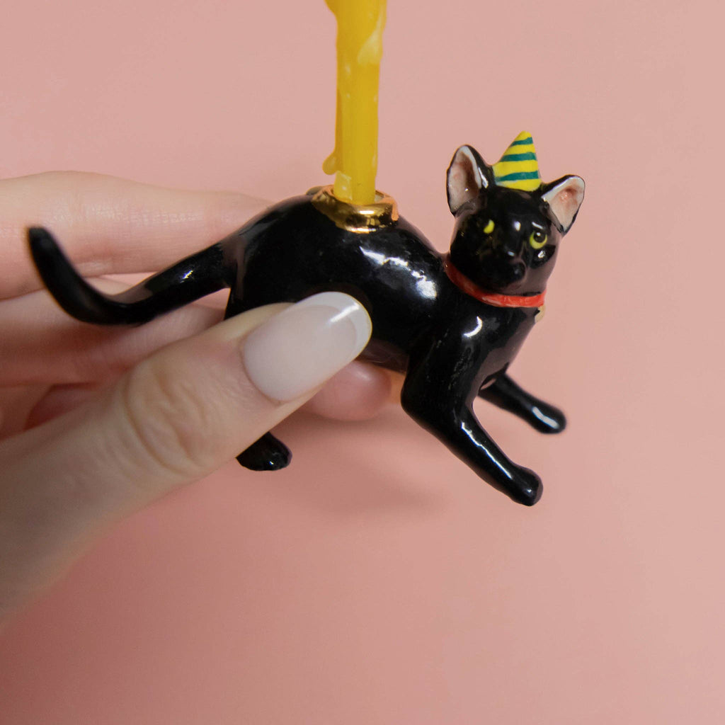 A hand holds the Black Cat Cake Topper, featuring a striped party hat and a yellow candle on its back. Ideal for cat-themed parties, it's set against a soft pink background.