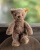 The Steiff Thommy Teddy Bear Brown Plush Toy, 8 inches, with Steiff’s signature yellow tag in its ear, sits on a wooden surface in front of a rustic fence and a softly blurred green and yellow background.