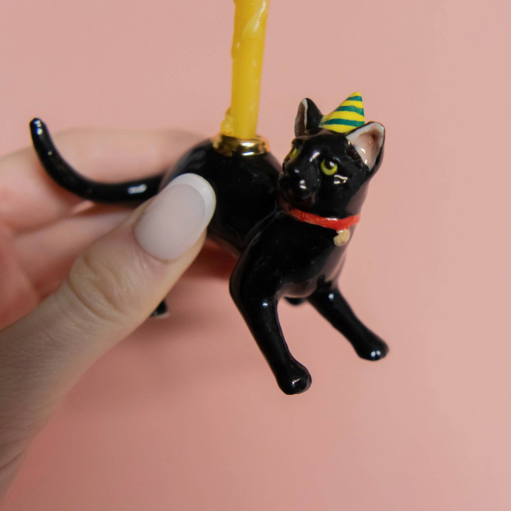A hand holds the Black Cat Cake Topper, featuring a lifelike black cat with a party hat and red collar. It also acts as a candle holder with a yellow candle on its back. Perfect for cat-themed parties, the soft pink background adds charm.