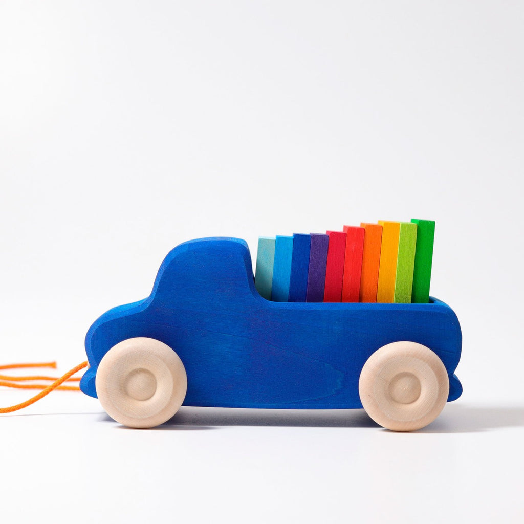 The Grimm's Pull Along Truck features natural wooden wheels, a non-toxic design, and an orange string for easy play. It carries rainbow-colored blocks stacked from red to green against a plain white background.
