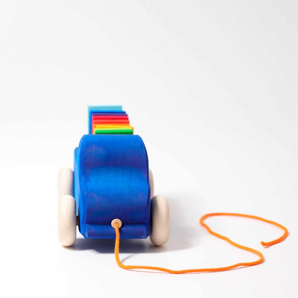 The Grimm's Pull Along Truck is a non-toxic blue wooden toy with four white wheels and an orange string. It features rainbow-colored blocks in red, yellow, and green stacked on top, resembling a charming truck pull toy. The product is set against a plain white background, perfect for kids.