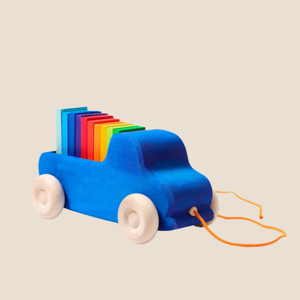 The Grimm's Pull Along Truck is a blue wooden toy with beige wheels and an orange string, featuring rainbow-colored blocks in green, blue, red, yellow, and pink stacked vertically. Crafted from non-toxic materials, it rests on a plain white background.