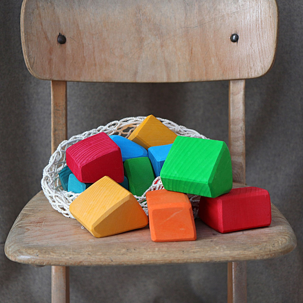 A wooden chair holds a net bag filled with vibrant Grimm's Colored Waldorf Blocks in red, green, blue, orange, and yellow. The blocks are stained with a non-toxic water-based color, ideal for open-ended indoor play against a plain background.