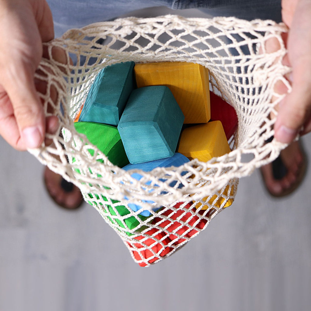A person standing on a light gray surface in sandals holds a net bag filled with Grimm's Colored Waldorf Blocks, stained with non-toxic, water-based colors, ready for open-ended play.