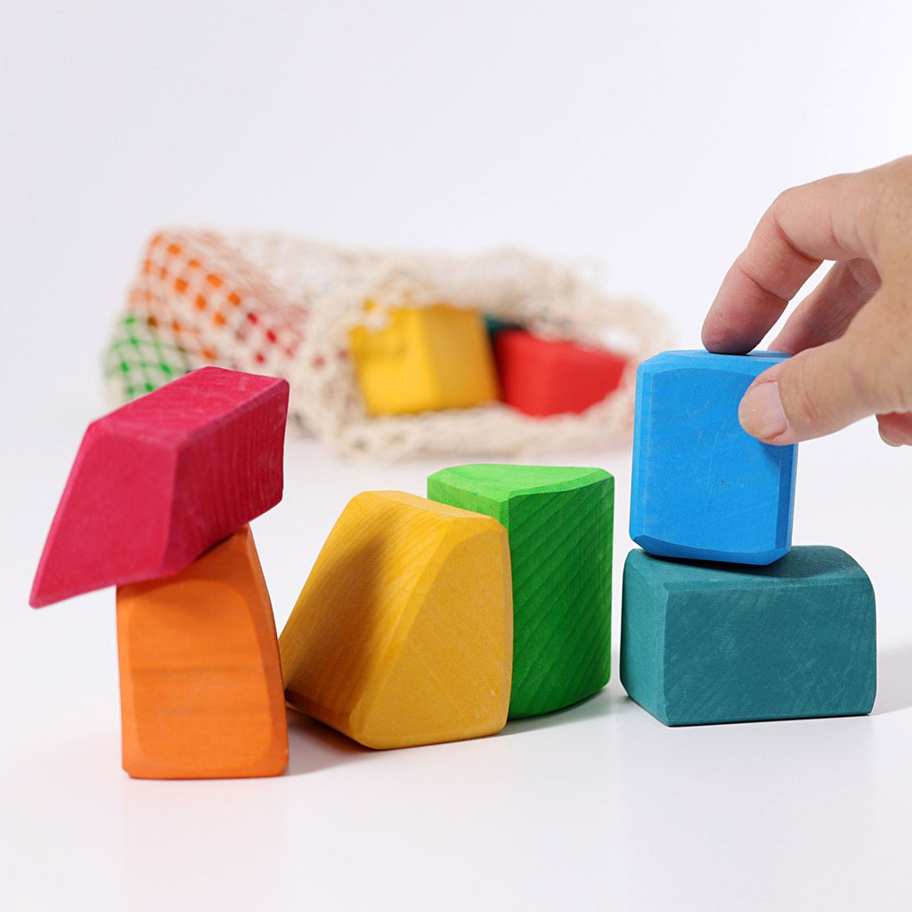 A hand stacks Grimm's Colored Waldorf Blocks in red, orange, yellow, green, and blue. These non-toxic blocks offer various shapes for open-ended play. A mesh bag with more awaits on a white surface to inspire creativity.