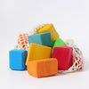 A vibrant collection of Grimm's Colored Waldorf Blocks in various shapes and hues, including red, blue, green, orange, and yellow, surrounds a mesh bag on a white background. These non-toxic blocks inspire open-ended play to spark endless creativity.