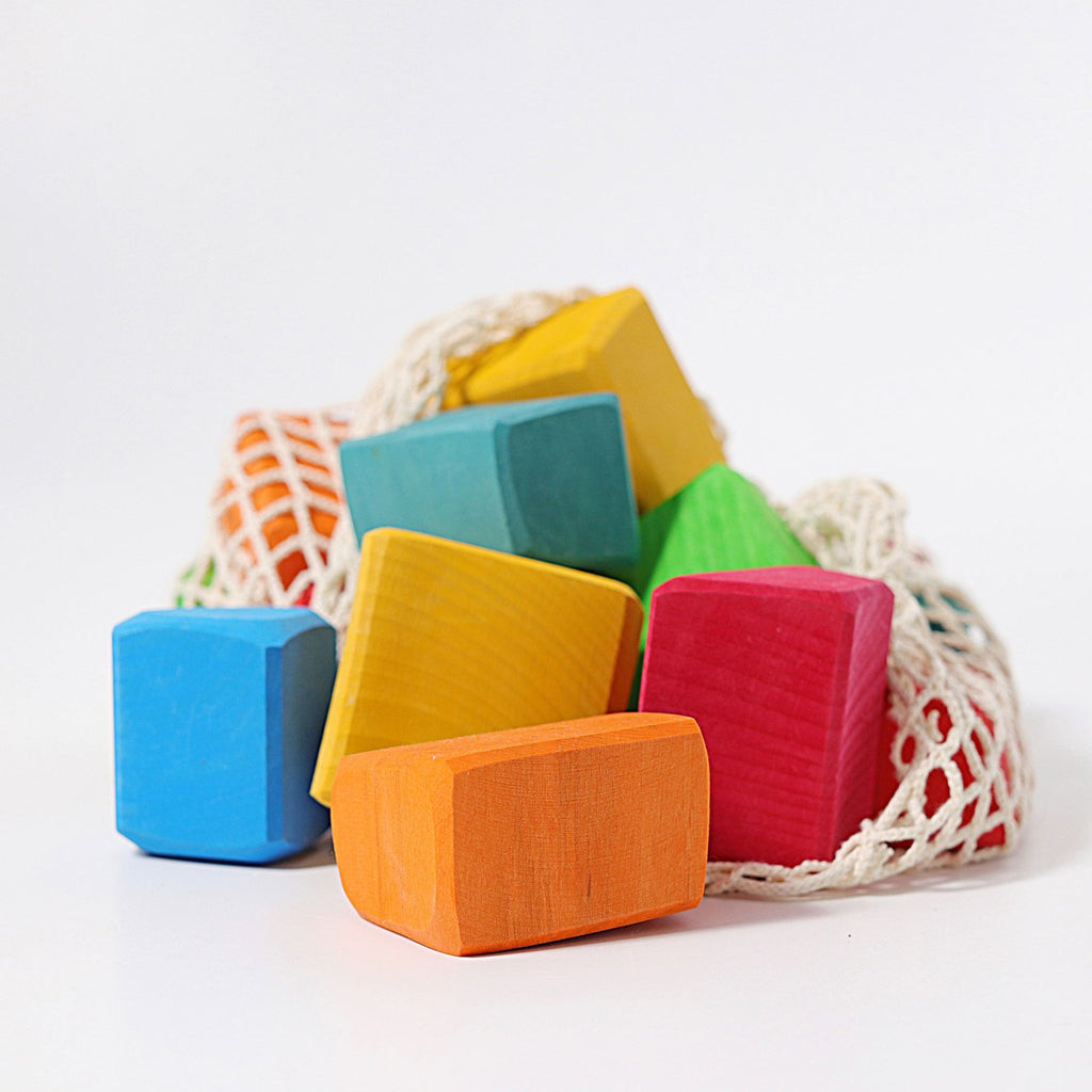 A vibrant collection of Grimm's Colored Waldorf Blocks in various shapes and hues, including red, blue, green, orange, and yellow, surrounds a mesh bag on a white background. These non-toxic blocks inspire open-ended play to spark endless creativity.