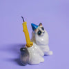 This charming Ragdoll Cat Cake Topper is a ceramic figurine with a fluffy tail, blue party hat, and yellow birthday candle against a lavender background—ideal for cat-themed parties.