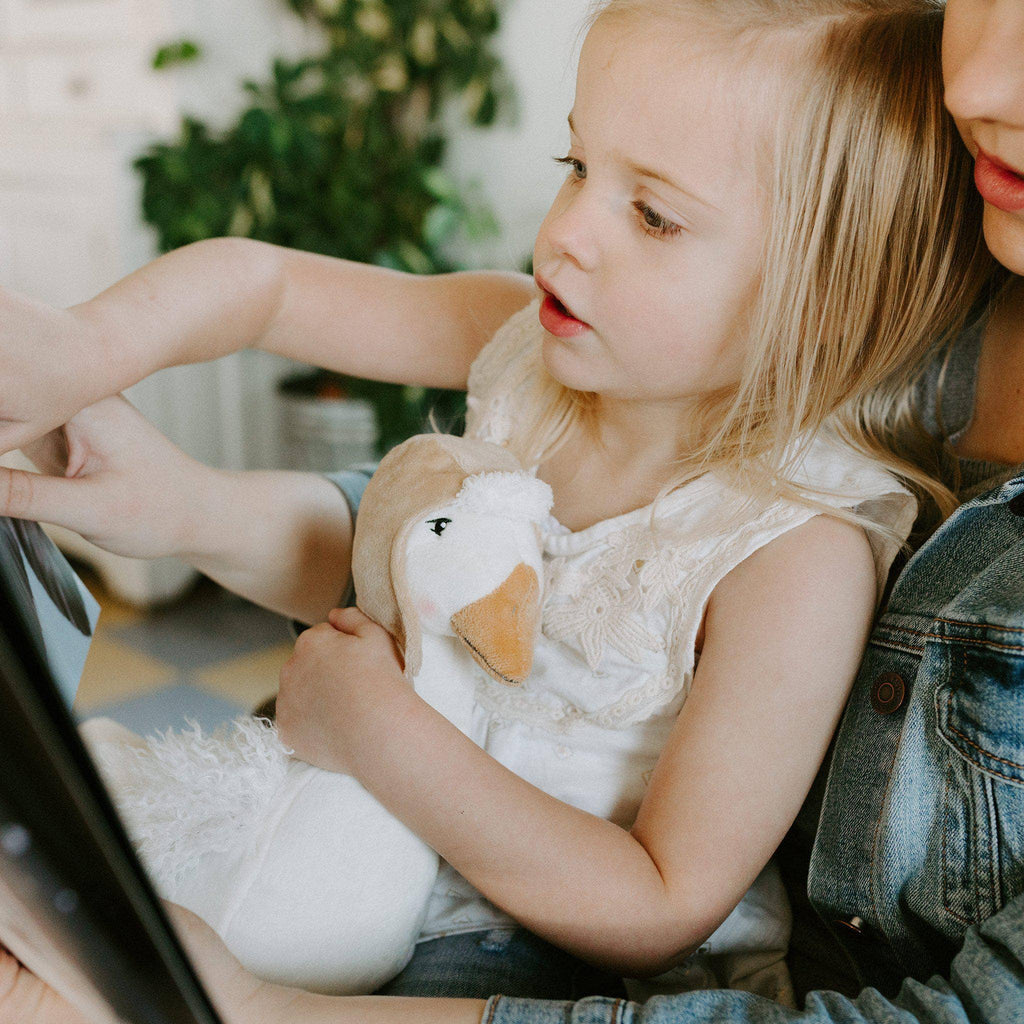 A young girl with blonde hair, wearing a white lace dress, sits on an adult's lap. She holds her beloved Avery the Aviator Snow Goose Stuffed Animal and looks intently at a tablet screen being pointed at by the adult. A potted plant is in the background.