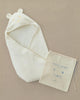 The Newborn Gift Set, featuring a cream-colored baby hooded swaddle with bear ears, crafted from organic cotton, comes with a matching drawstring bag labeled "hooded swaddle" and an animal face design. Elegantly displayed on a beige background, it makes the perfect baby gift.