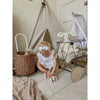 A baby in a white organic cotton outfit plays with a wooden car on a rug. Nearby are a wicker basket, toy cart with the Senger Naturwelt Stuffed Animal - White Bunny, and a small fabric teepee in the cozy, neutral-toned room.