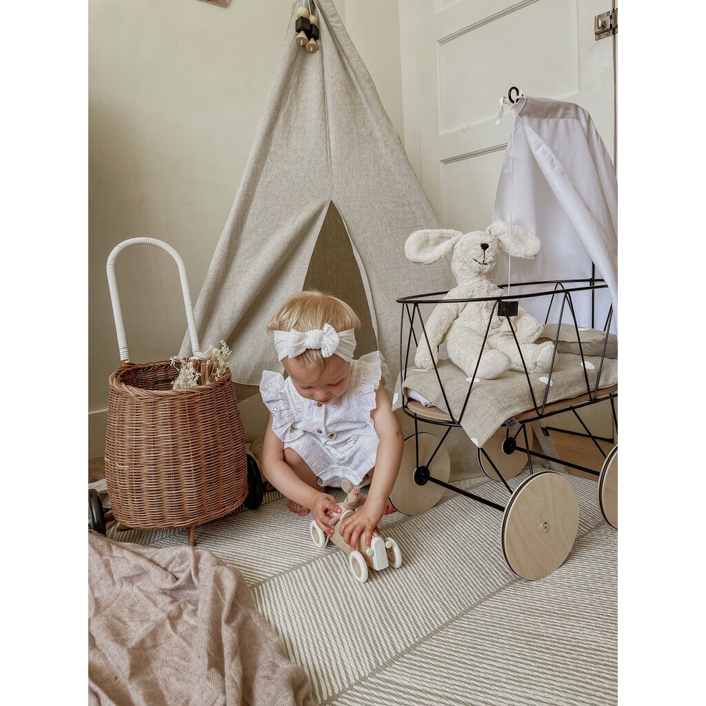 A baby in a white organic cotton outfit plays with a wooden car on a rug. Nearby are a wicker basket, toy cart with the Senger Naturwelt Stuffed Animal - White Bunny, and a small fabric teepee in the cozy, neutral-toned room.