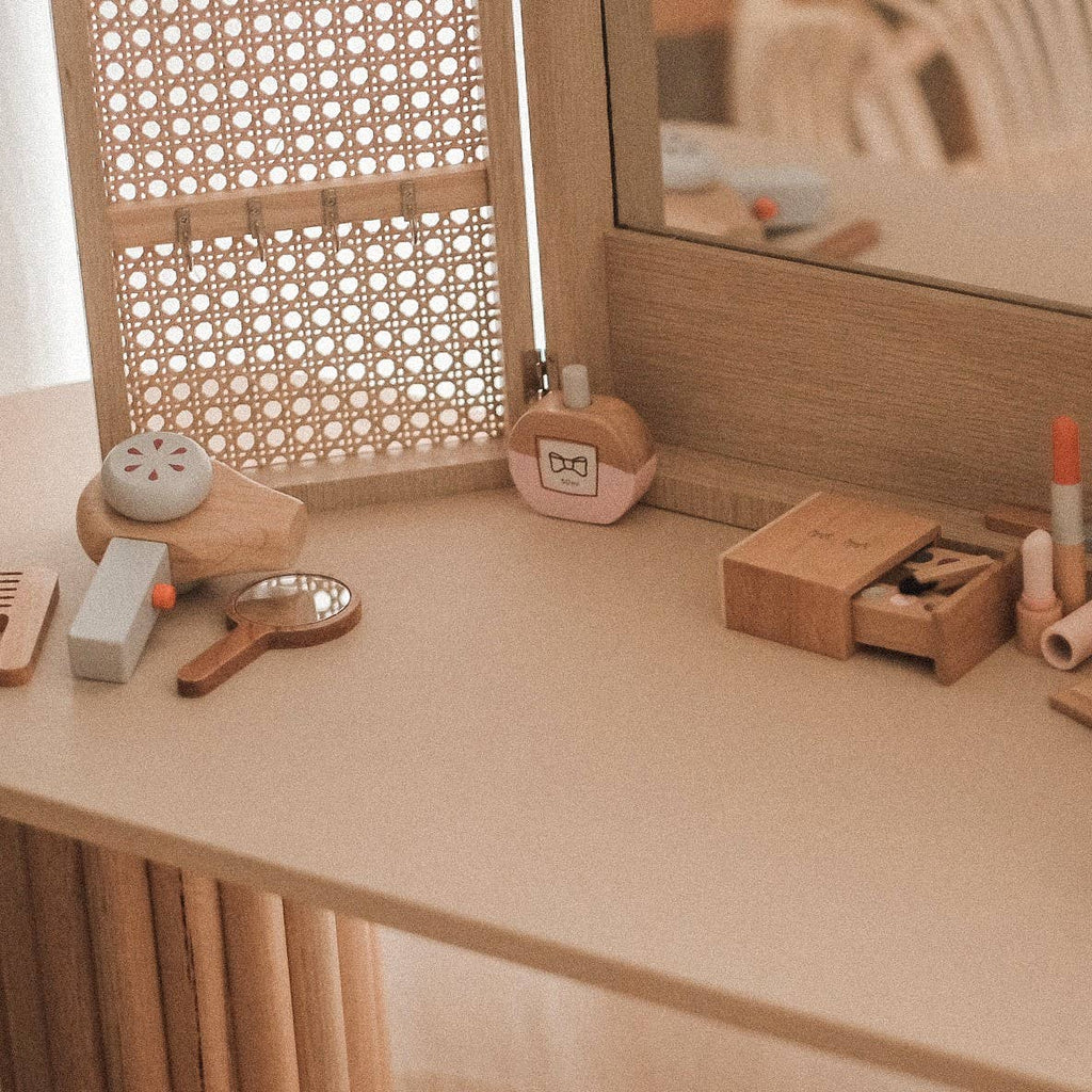The Wooden Beauty Kit is showcased on a table, including wooden toys like a mirror, comb, powder compact, lipstick, and eye shadow. Non-toxic paint ensures safe play. A woven screen and large mirror in the background enhance the cozy vanity setup.