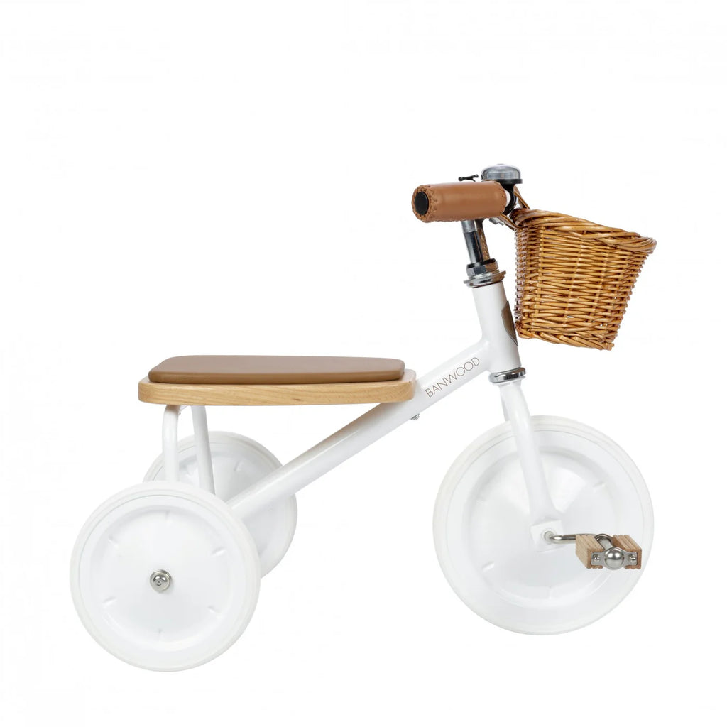 The Banwood Toddler Trike is a minimalist white tricycle with a brown seat and handlebars, featuring smooth white wheels and a front wicker basket for toddlers.