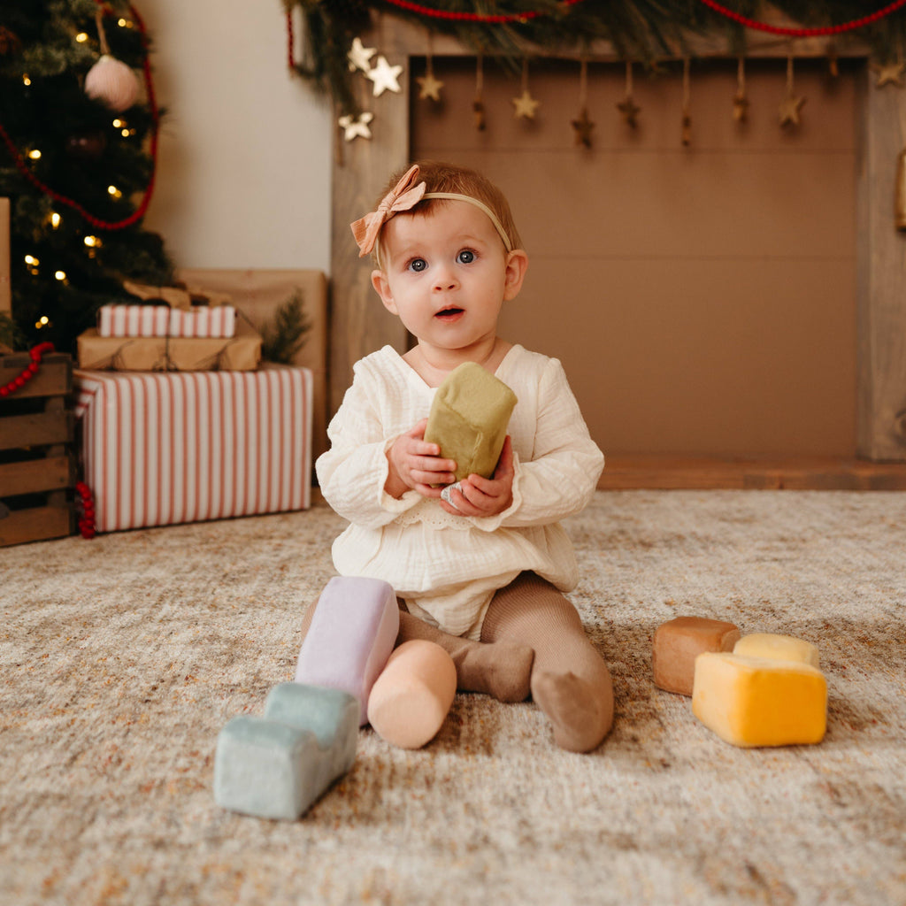 A toddler with a bow headband sits on a rug holding a large green Soft Toy Block, while other colorful Soft Toy Blocks are scattered nearby. Wrapped presents and a decorated Christmas tree are in the background.