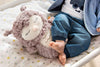 A baby in blue pants sits on star-patterned sheets, resting bare feet against a Steiff Ollie Owl Stuffed Animal with closed eyes and a soft, premium-grade plush texture.
