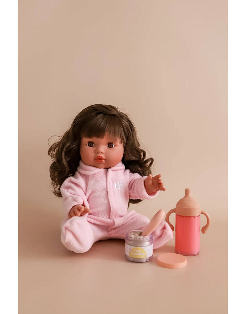 The My Tiny Birthday Set by Tiny Harlow features a doll with long brown hair seated on a beige surface, dressed in a pink onesie. In front of her are a pink baby bottle and a mini container styled like a birthday cake jar, evoking realistic play. The backdrop is plain light brown.