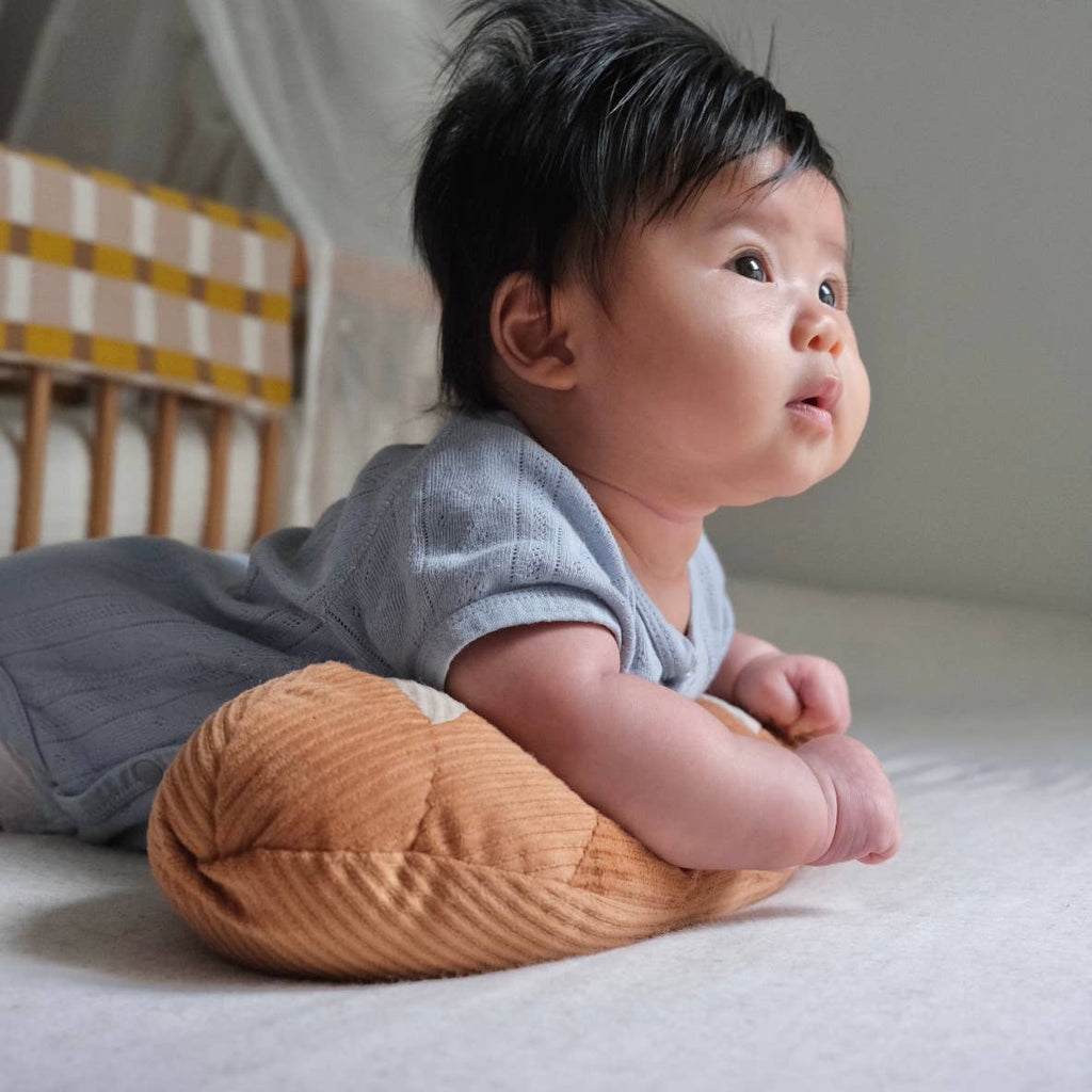A dark-haired baby in a blue outfit lies on their stomach, supported by the Tummy Time Baguette pillow, gazing upward with curiosity. Behind them is a crib featuring a yellow and white checkered pattern.