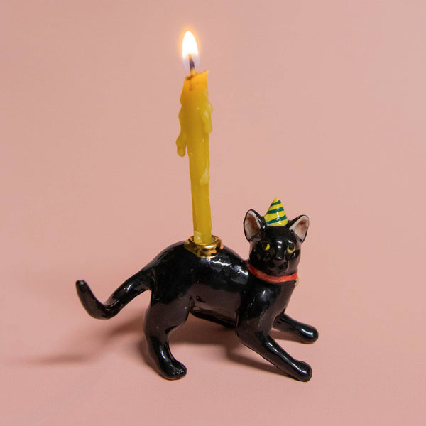 The Black Cat Cake Topper showcases a ceramic black cat with lifelike details, featuring a red collar, green-and-yellow striped party hat, and a yellow candle on its back. It sits against a soft peach background, making it ideal for cat-themed parties.