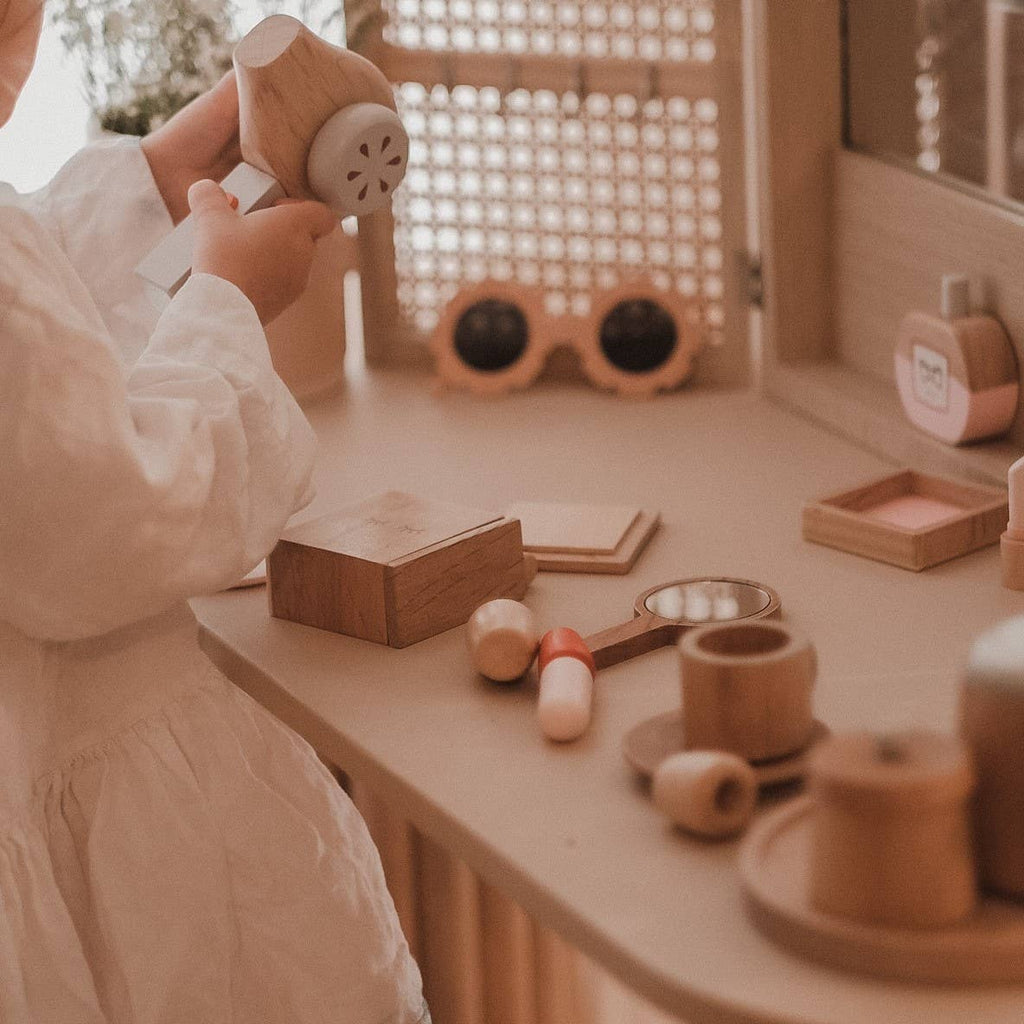 In a cozy room, a child in a white shirt plays with the timeless Wooden Beauty Kit. The table is scattered with wooden toys and objects as soft light filters through the window, illuminating the warm earthy tones of the setting.