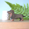 The Handmade Wooden Dachshund Dog figurine, crafted from sustainable hardwood and finished with non-toxic plant-based stains, features a simple carved design. It sits on a wooden surface beside a white vase and green fern leaves.