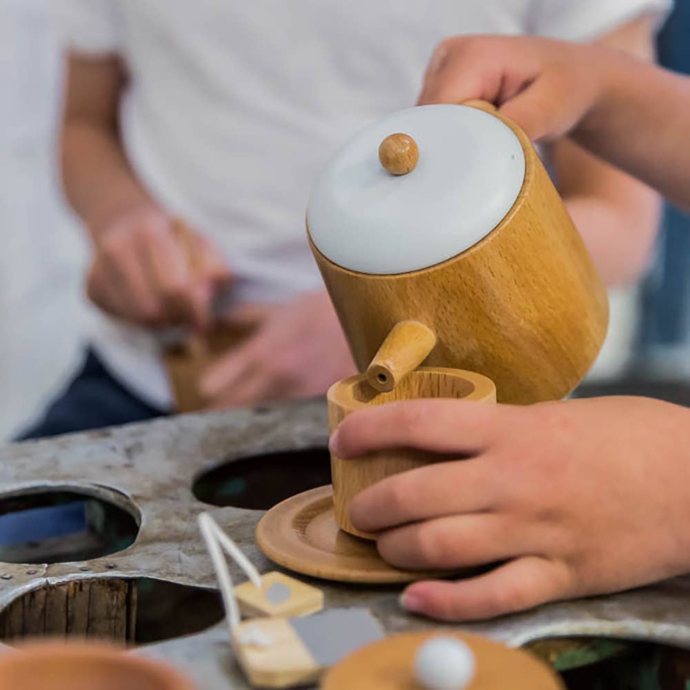 Hands pour tea from a Wooden Tea Set into a matching cup on a saucer, while another person in the background holds a cup. The scene sits atop a designer coffee table with circular cutouts, creating a casual yet cozy ambiance.