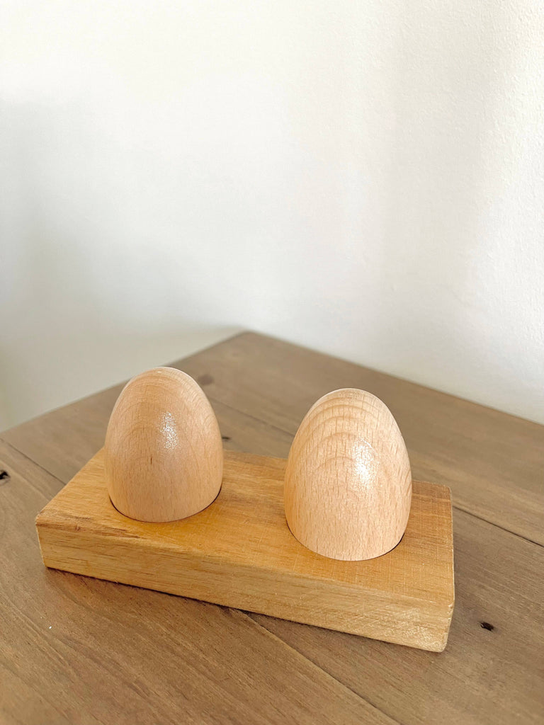The Wooden Egg Shakers with Stand features two smooth, dome-shaped egg shakers crafted from natural wood, displayed on a light wood base—ideal for supporting developmental milestones.