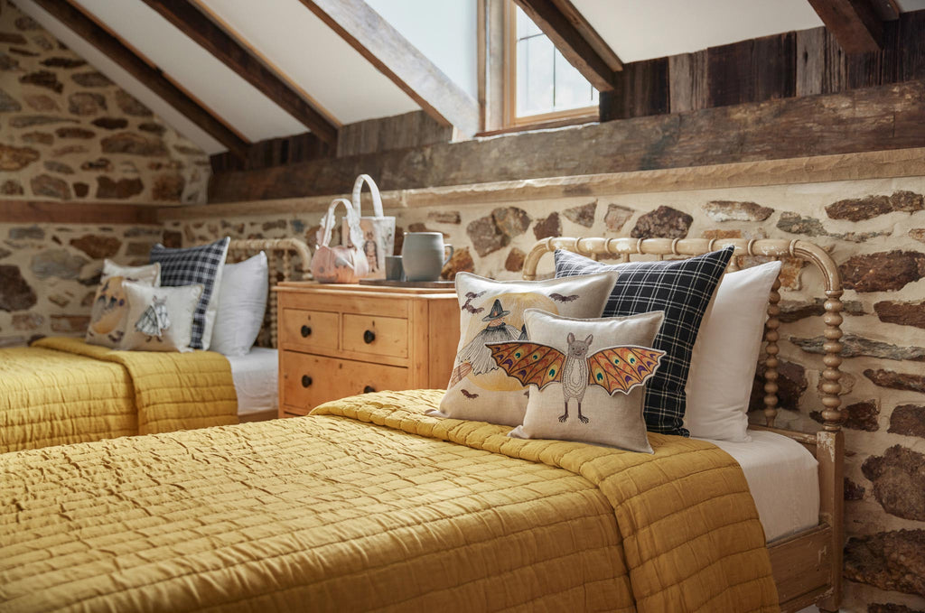 Cozy rustic bedroom featuring two twin beds with yellow quilts and decorative pillows, including a Bat Wing Surprise Pillow, set against a stone wall, with exposed wooden beams and a bright window.