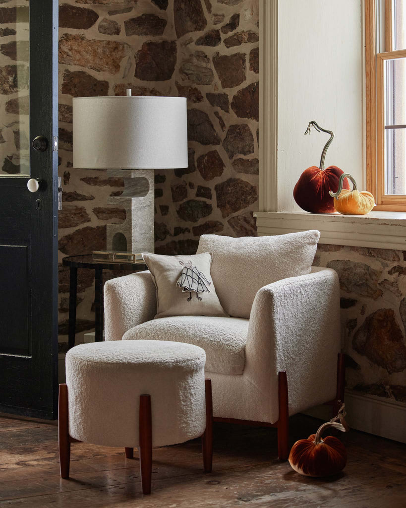 A cozy interior featuring a plush white armchair with a Bat Wing Surprise Pillow resting next to a window with autumn pumpkins on the sill, all against an old stone wall.