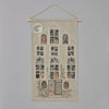Introducing the Haunted House Advent Calendar: a captivating tapestry depicting a haunted house with 16 windows, each revealing spooky characters such as ghosts and skeletons. This Halloween advent calendar is beautifully adorned with pumpkins and plants, and features a full moon in the top left corner. It hangs elegantly on a wooden dowel with a string.