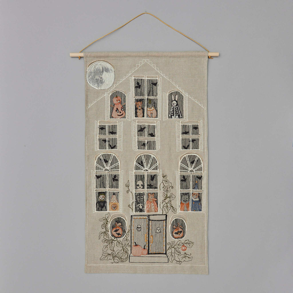 Introducing the Haunted House Advent Calendar: a captivating tapestry depicting a haunted house with 16 windows, each revealing spooky characters such as ghosts and skeletons. This Halloween advent calendar is beautifully adorned with pumpkins and plants, and features a full moon in the top left corner. It hangs elegantly on a wooden dowel with a string.