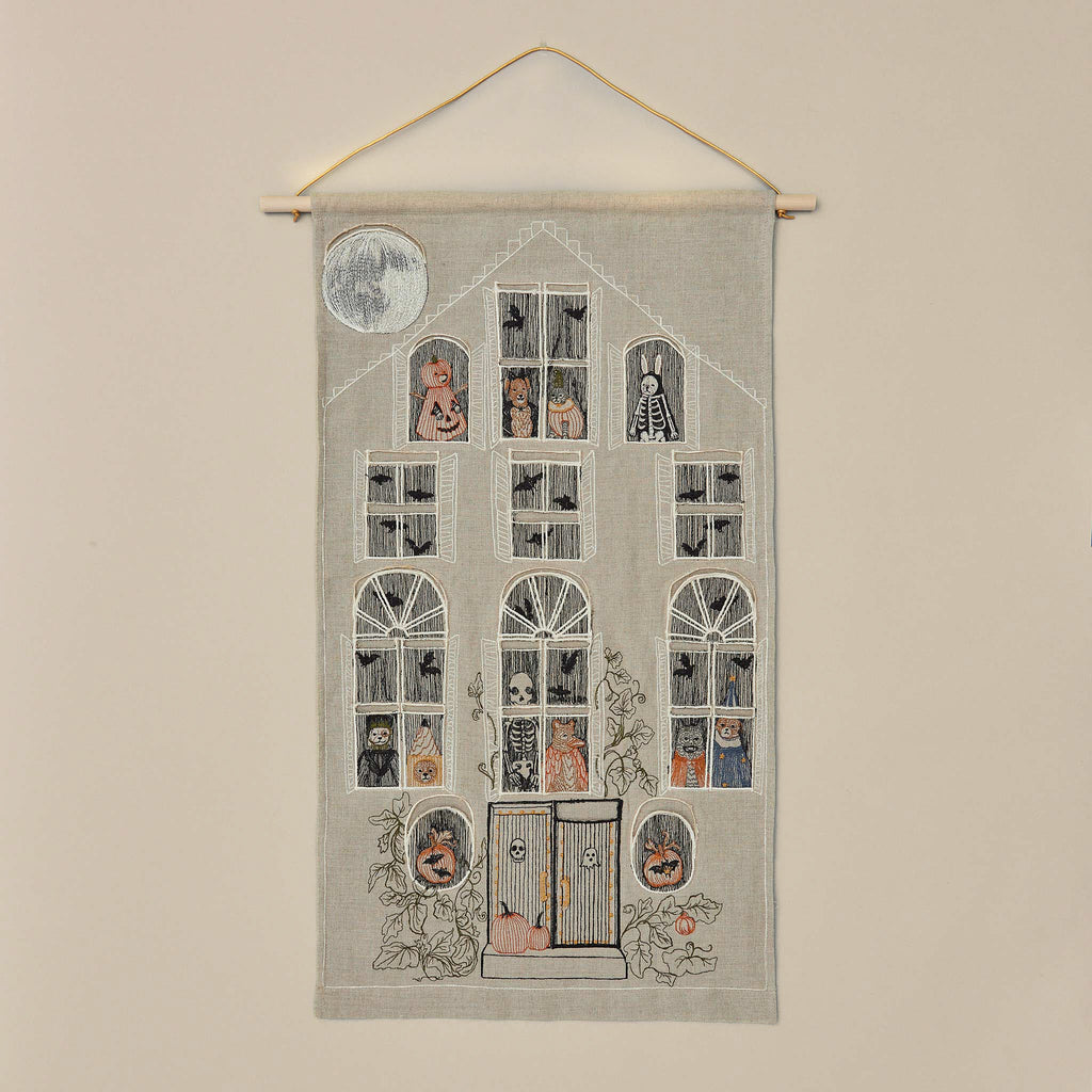 The Haunted House Advent Calendar is an embroidered fabric art piece designed as a multi-story haunted house. It features various Halloween characters, such as skeletons and pumpkins, visible through the windows. The house boasts intricate Halloween-themed decorations and a full moon glowing in the top left corner.