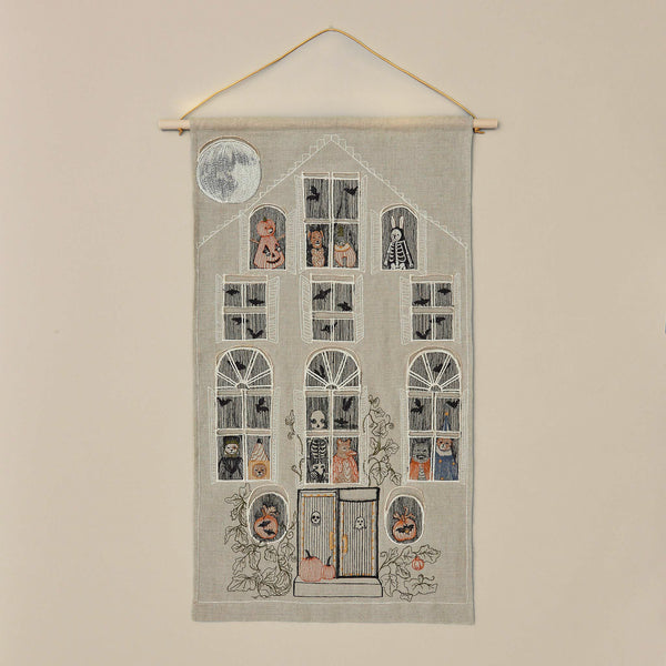 The Haunted House Advent Calendar is an embroidered fabric art piece designed as a multi-story haunted house. It features various Halloween characters, such as skeletons and pumpkins, visible through the windows. The house boasts intricate Halloween-themed decorations and a full moon glowing in the top left corner.