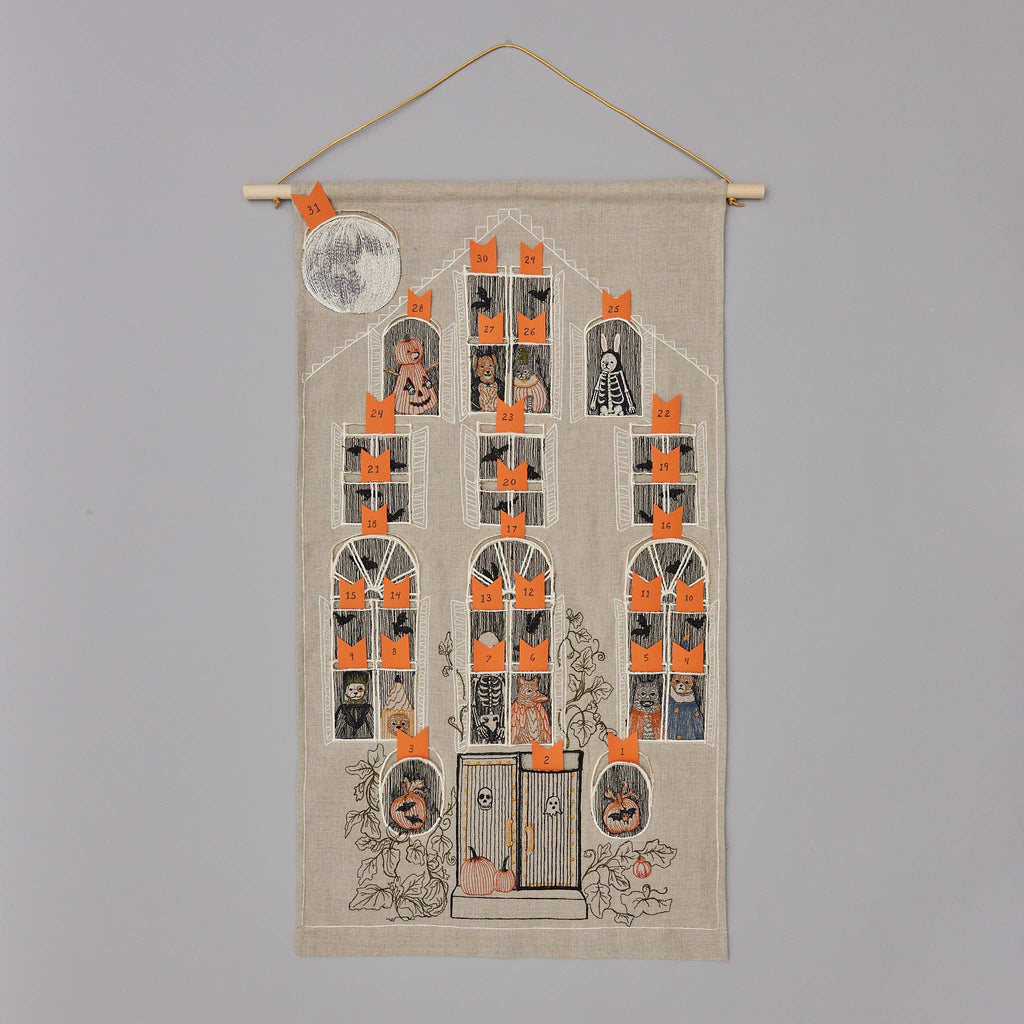 The Haunted House Advent Calendar is a fabric wall hanging featuring a spooky haunted house with 25 orange, numbered windows for a Halloween countdown. The house is adorned with ghostly figures, pumpkins, skeletons, and other eerie elements. A full moon illuminates the scene, revealing both tricks and treats as you count down to Halloween.