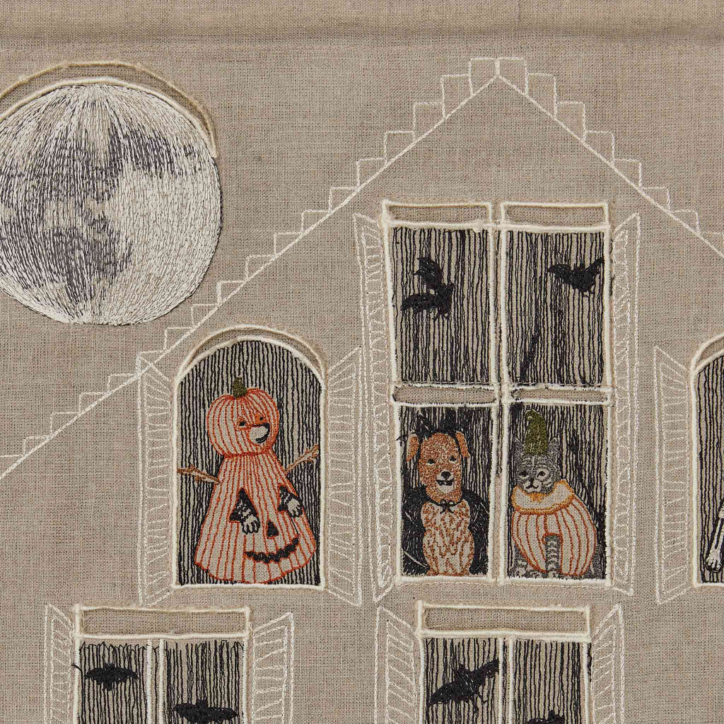 This Haunted House Advent Calendar features embroidered artwork of a spooky house under a full moon. The house has four windows showcasing different creatures: two display figures in costumes, while the other two reveal cats. Black bats flutter both in and out of the windows, enhancing the Halloween tricks and treats atmosphere.
