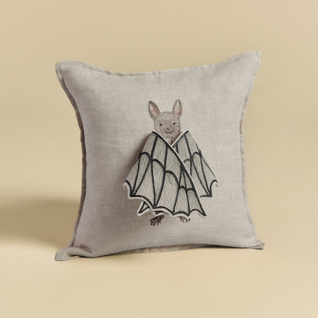 A Bat Wing Surprise Pillow with a beige background featuring an embroidered design of a bat with butterfly wings, showcasing detailed stitching on a neutral-toned fabric.