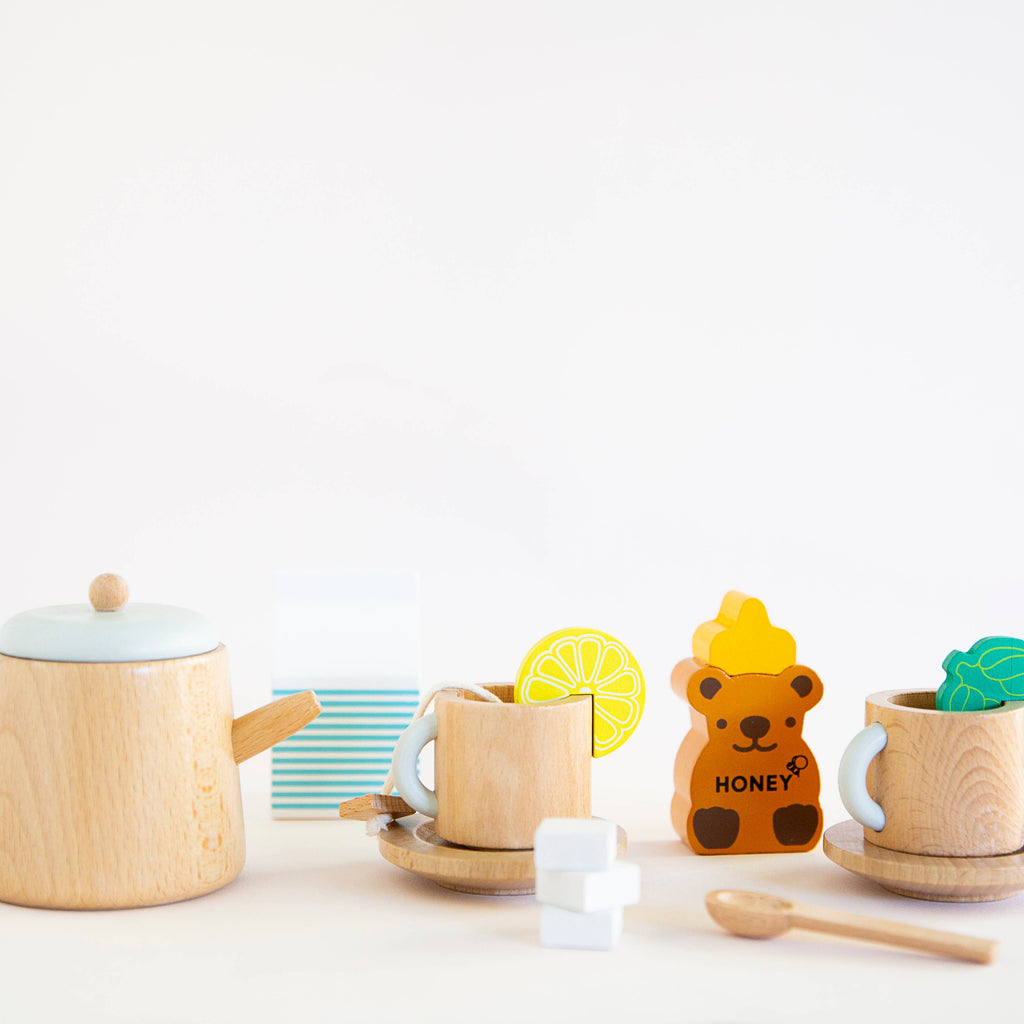 The Wooden Tea Set is ideal for a cozy play space, featuring a teapot, lemon-adorned cups, sugar cubes, a bear-shaped honey jar, a wooden spoon, and milk carton.