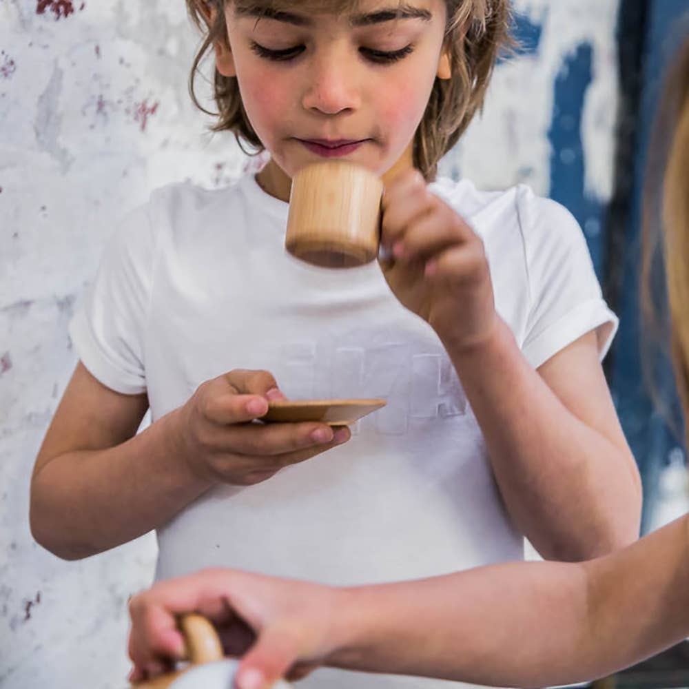 A child in a white shirt sips from the Wooden Tea Set's cup and saucer while another hand pours liquid, creating a scene akin to designer coffee table book pages set against a textured white and blue wall.