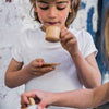A child in a white shirt sips from the Wooden Tea Set's cup and saucer while another hand pours liquid, creating a scene akin to designer coffee table book pages set against a textured white and blue wall.