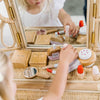 A child engages in imaginative play with the Wooden Beauty Kit on a wicker vanity, featuring a lipstick, compact, and non-toxic paint. Their reflection comes alive in the mirror as they explore each miniature cosmetic piece.