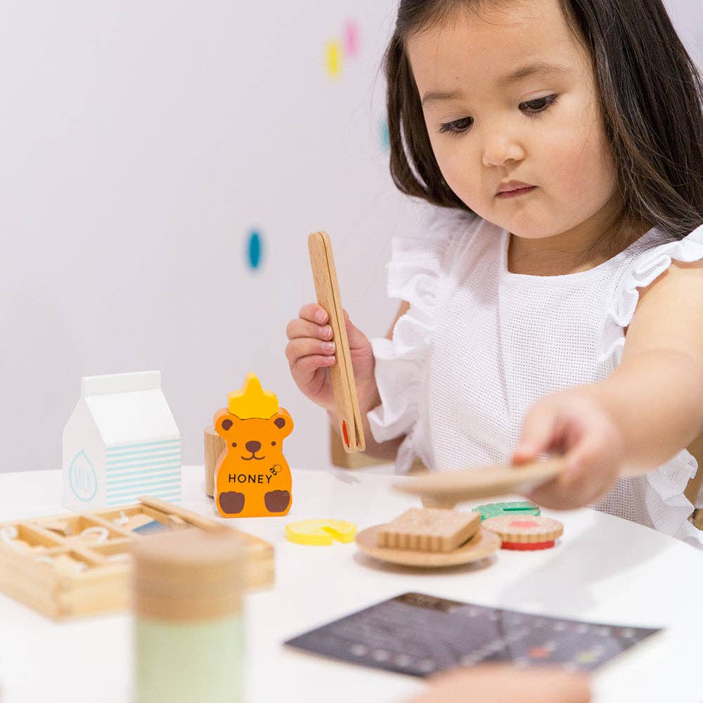 In her imaginative play space, a young child with long dark hair uses tongs to pick up toy food from the Wooden Tea Set. Nearby are a honey bear container and wooden bread, and an organic Matcha Tea box. She is focused, wearing a white top.