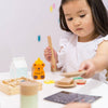 In her imaginative play space, a young child with long dark hair uses tongs to pick up toy food from the Wooden Tea Set. Nearby are a honey bear container and wooden bread, and an organic Matcha Tea box. She is focused, wearing a white top.