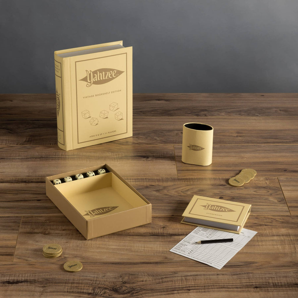 A Yahtzee Vintage Bookshelf Edition board game set with cream dice, scorecards, pen, tokens, cup, and a book-shaped box is elegantly displayed on a wooden table.