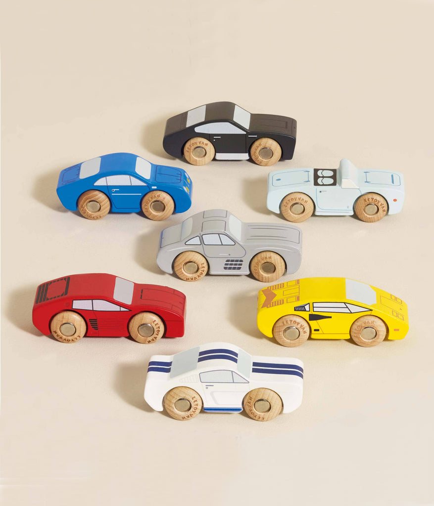 A set of seven Classic Wooden Sports Cars, made from FSC-certified wood in blue, black, gray, light blue, red, white, and yellow with unique details and wooden wheels is displayed on a beige surface. These colorful cars are train track compatible for endless fun.