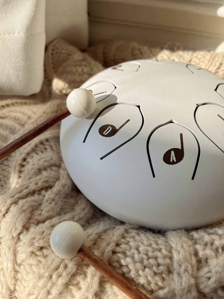 A Steel Tongue Drum, a unique musical instrument, with wooden mallets rests on a knitted cream blanket. Sunlight casts soft shadows, highlighting the "D" and "A" notes on its white steel surface.
