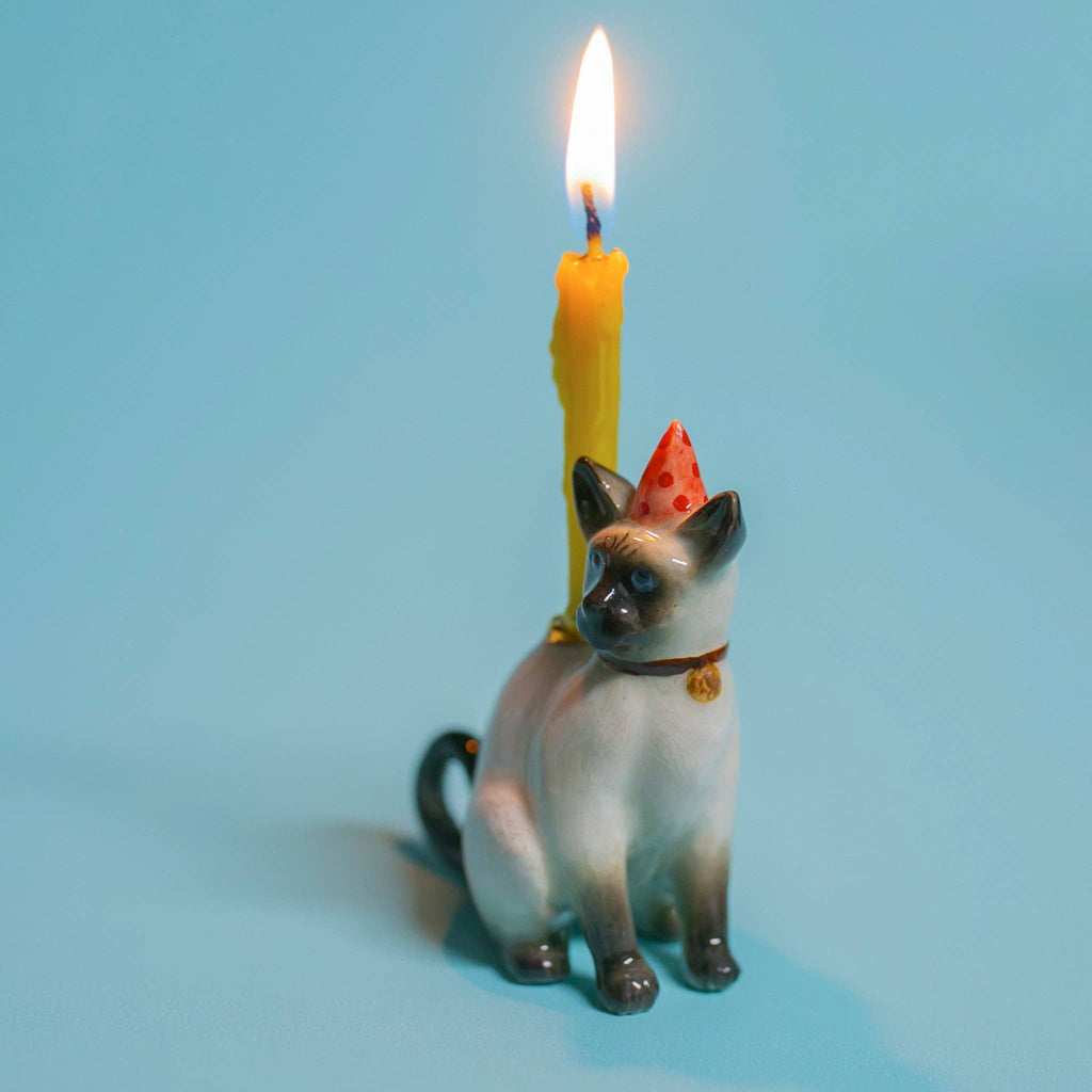 The Siamese Cat Cake Topper is a ceramic figurine with a party hat and lit yellow candle on its back, set against a light blue background, ideal for cat-themed parties.